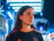 IMG_5238 | AllezPaillade.com Woman in a dark sports jersey with orange stripes and a circular chest logo, standing in a blue-lit store or showroom, looking to the right.