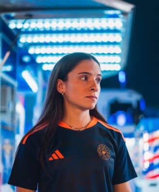 IMG_5238 | AllezPaillade.com Woman in a dark sports jersey with orange stripes and a circular chest logo, standing in a blue-lit store or showroom, looking to the right.