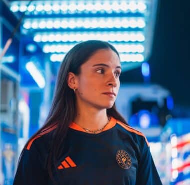 Woman in a dark sports jersey with orange stripes and a circular chest logo, standing in a blue-lit store or showroom, looking to the right.