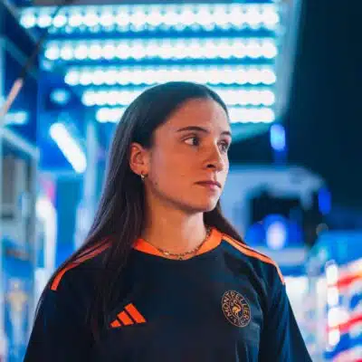Woman in a dark sports jersey with orange stripes and a circular chest logo, standing in a blue-lit store or showroom, looking to the right.