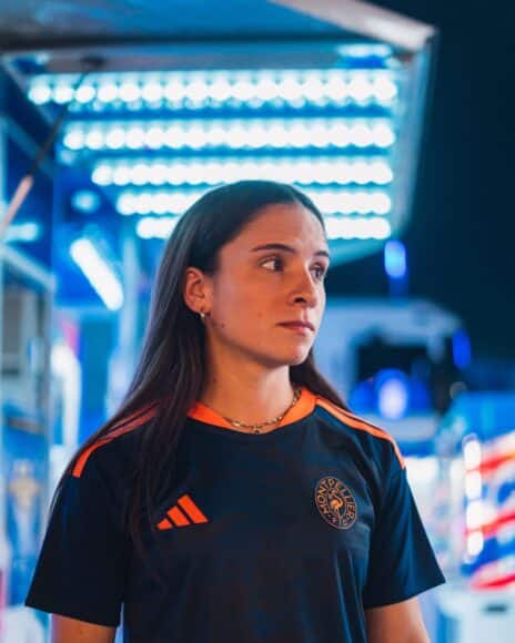 Woman in a dark sports jersey with orange stripes and a circular chest logo, standing in a blue-lit store or showroom, looking to the right.