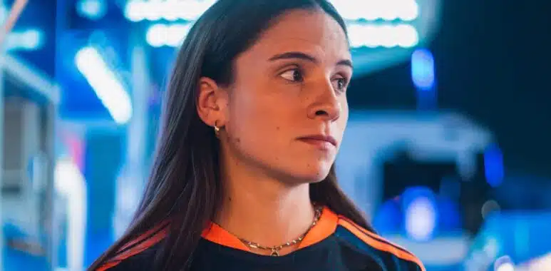 Woman in a dark sports jersey with orange stripes and a circular chest logo, standing in a blue-lit store or showroom, looking to the right.