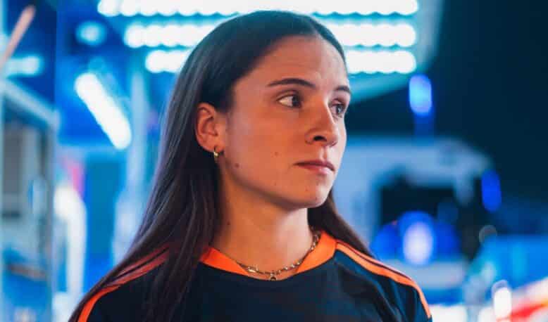 IMG_5238 | AllezPaillade.com Woman in a dark sports jersey with orange stripes and a circular chest logo, standing in a blue-lit store or showroom, looking to the right.