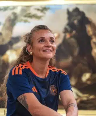 Woman with long hair in a blue and orange sports jersey smiles while standing in front of a large framed painting in an art gallery.