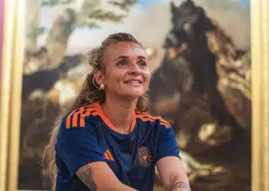 Woman with long hair in a blue and orange sports jersey smiles while standing in front of a large framed painting in an art gallery.