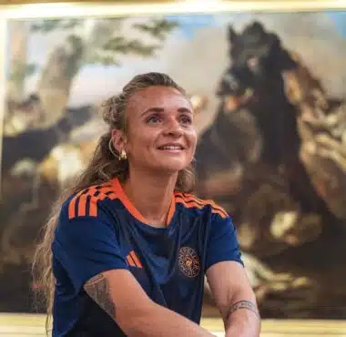 Woman with long hair in a blue and orange sports jersey smiles while standing in front of a large framed painting in an art gallery.