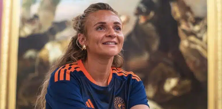 Screenshot | AllezPaillade.com Woman with long hair in a blue and orange sports jersey smiles while standing in front of a large framed painting in an art gallery.