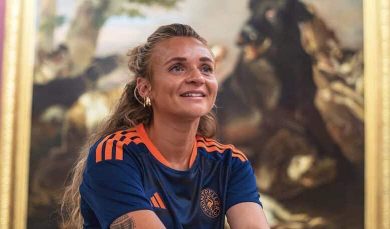 Screenshot | AllezPaillade.com Woman with long hair in a blue and orange sports jersey smiles while standing in front of a large framed painting in an art gallery.