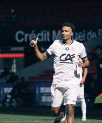 Screenshot | AllezPaillade.com Soccer player in a white jersey and shorts runs on the field, giving a thumbs-up during a match.