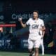 Soccer player in a white jersey and shorts runs on the field, giving a thumbs-up during a match.
