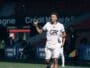 Soccer player in a white jersey and shorts runs on the field, giving a thumbs-up during a match.