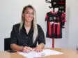 Woman seated at a table signing documents with a pen, a black-and-red sports jersey on a stand in the background.