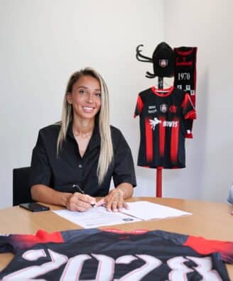 Woman seated at a table signing documents with a pen, a black-and-red sports jersey on a stand in the background.