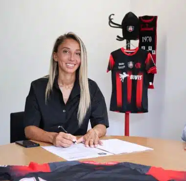 Woman seated at a table signing documents with a pen, a black-and-red sports jersey on a stand in the background.