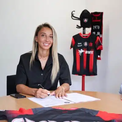 Woman seated at a table signing documents with a pen, a black-and-red sports jersey on a stand in the background.