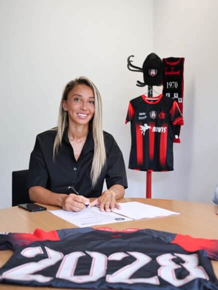 Woman seated at a table signing documents with a pen, a black-and-red sports jersey on a stand in the background.