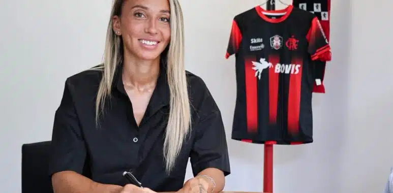 IMG_5280 | AllezPaillade.com Woman seated at a table signing documents with a pen, a black-and-red sports jersey on a stand in the background.