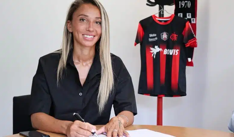 Woman seated at a table signing documents with a pen, a black-and-red sports jersey on a stand in the background.