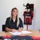 Woman seated at a table signing documents with a pen, a black-and-red sports jersey on a stand in the background.