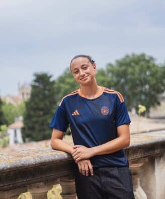 Teenager in a navy and orange soccer jersey leaning on a stone railing outdoors with trees in the background.