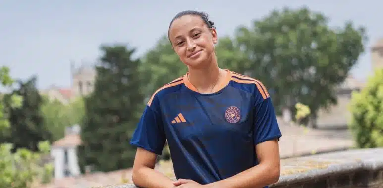 Screenshot | AllezPaillade.com Teenager in a navy and orange soccer jersey leaning on a stone railing outdoors with trees in the background.