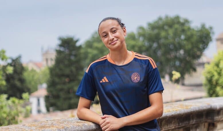 Teenager in a navy and orange soccer jersey leaning on a stone railing outdoors with trees in the background.