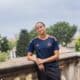 Teenager in a navy and orange soccer jersey leaning on a stone railing outdoors with trees in the background.