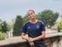 Teenager in a navy and orange soccer jersey leaning on a stone railing outdoors with trees in the background.