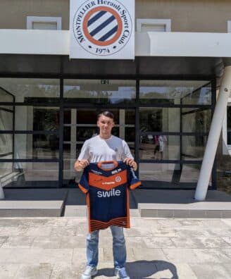 Young man standing outside a sports club entrance, holding a blue and orange team jersey.