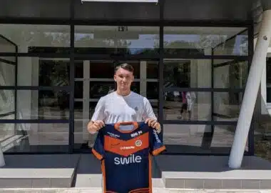 Young man standing outside a sports club entrance, holding a blue and orange team jersey.
