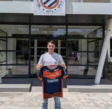 Young man standing outside a sports club entrance, holding a blue and orange team jersey.