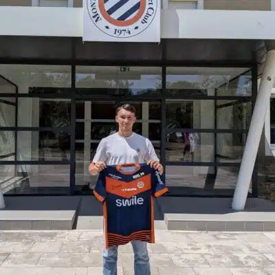 Young man standing outside a sports club entrance, holding a blue and orange team jersey.
