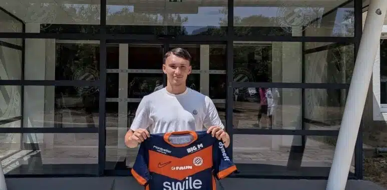 Young man standing outside a sports club entrance, holding a blue and orange team jersey.
