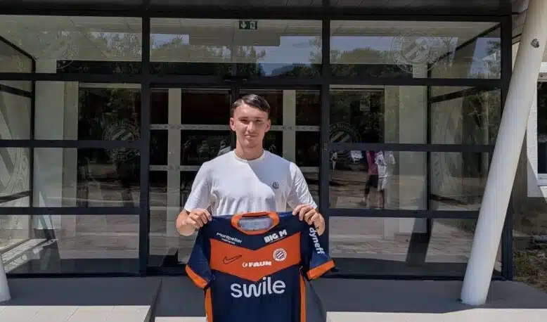 Young man standing outside a sports club entrance, holding a blue and orange team jersey.