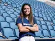 Woman with long brown hair standing in empty blue stadium seats, arms crossed and smiling at the camera.