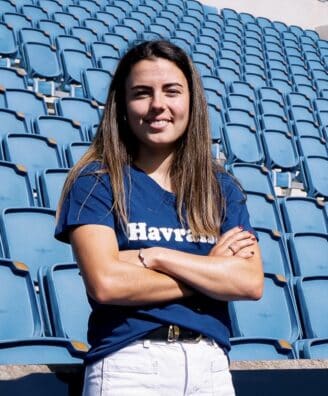 Screenshot | AllezPaillade.com Woman with long brown hair standing in empty blue stadium seats, arms crossed and smiling at the camera.