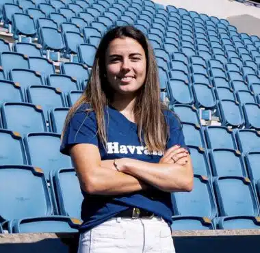 Woman with long brown hair standing in empty blue stadium seats, arms crossed and smiling at the camera.