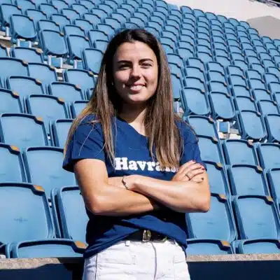 Woman with long brown hair standing in empty blue stadium seats, arms crossed and smiling at the camera.