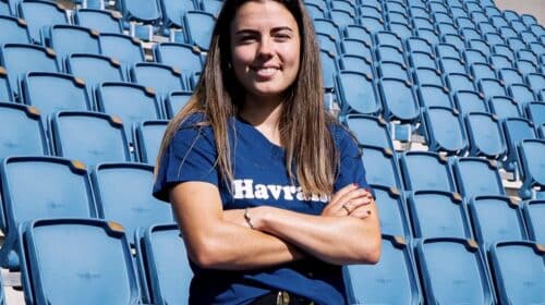 Woman with long brown hair standing in empty blue stadium seats, arms crossed and smiling at the camera.