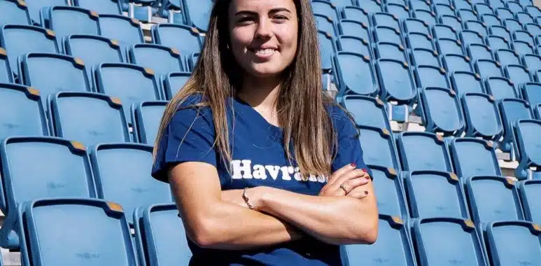 Screenshot | AllezPaillade.com Woman with long brown hair standing in empty blue stadium seats, arms crossed and smiling at the camera.