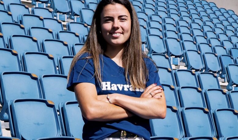 Woman with long brown hair standing in empty blue stadium seats, arms crossed and smiling at the camera.