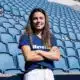 Woman with long brown hair standing in empty blue stadium seats, arms crossed and smiling at the camera.