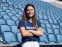Woman with long brown hair standing in empty blue stadium seats, arms crossed and smiling at the camera.