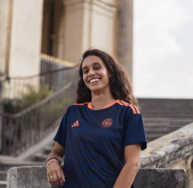Smiling woman in a navy blue and orange sports jersey standing by stone stairs and railing outdoors.