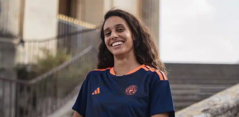 Screenshot | AllezPaillade.com Smiling woman in a navy blue and orange sports jersey standing by stone stairs and railing outdoors.