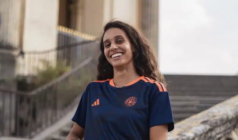 Smiling woman in a navy blue and orange sports jersey standing by stone stairs and railing outdoors.