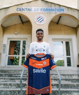 Young man stands on concrete steps outside a training center, holding a blue and orange football jersey toward the camera.