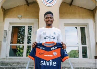 Young man stands on concrete steps outside a training center, holding a blue and orange football jersey toward the camera.