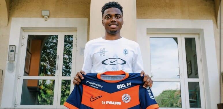 Young man stands on concrete steps outside a training center, holding a blue and orange football jersey toward the camera.