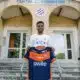 Young man stands on concrete steps outside a training center, holding a blue and orange football jersey toward the camera.